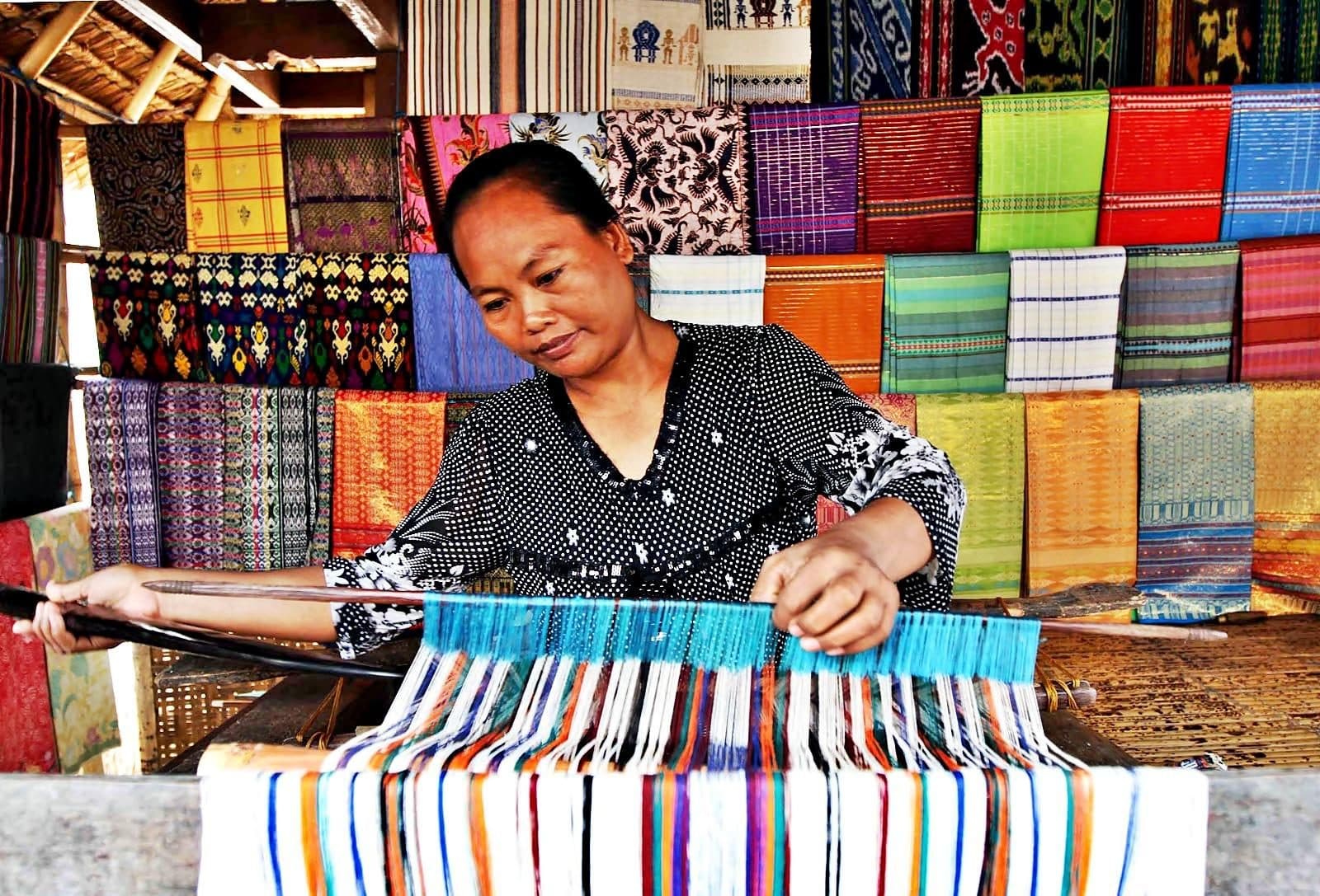 A weaver at a traditional loom with colorful Sasak textiles behind her